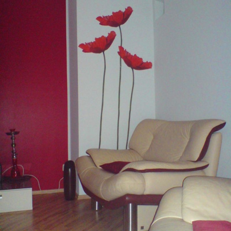 Wohnzimmer in der eine Wandabteil in dunkelrot mit Pinkanteil gestrichen wurde, de restlichen Wand weiß und ein schmales Stück Wand wurde mit 3 riesigen Mohnblumen, die vom Boden bis zur Decke reichen, in der Farbe der roten Wand verziert.